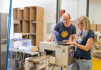 Das Bild zeigt zwei Mitarbeiter, die mit einem Laptop bei einer Maschine mit Förderband stehen.  | © SONNENTOR